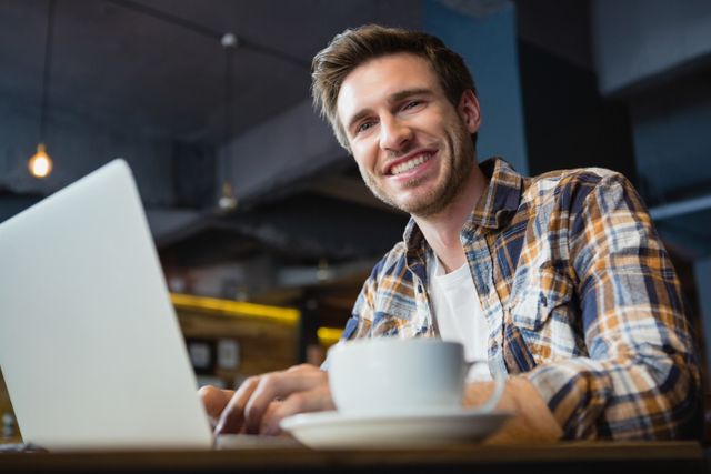 Portrait of happy young man using laptop while having coffee in cafÃ©