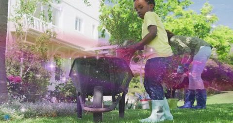 Child planting potted shrub with wheelbarrow on sunny suburban lawn wearing yellow boots