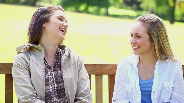 Young women engage in lively conversation on park bench, ideal for concepts related to friendship, healthy social interactions, outdoor leisure activities, or mental well-being. Useful for promotional materials, articles, or blog posts focusing on positive social experiences and youthful connections.