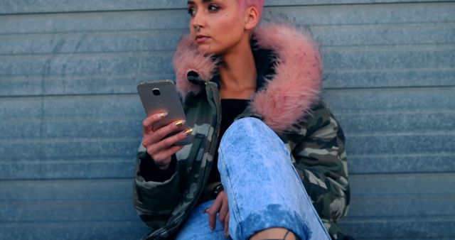 This image features a fashionable young woman with pink hair, using a smartphone while sitting casually in an urban environment. She is wearing a camouflage jacket with a pink hood trim and ripped jeans. This photo can be used to represent fashion, modern lifestyles, technology use, or urban youth culture. Ideal for blogs, social media content, advertisements, and articles related to trends, technology, or lifestyle.