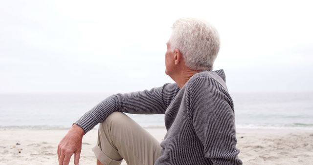 Senior man is sitting on a sandy beach wearing a grey sweater and beige pants, gazing thoughtfully at the horizon over the ocean. This scene can be used in retirement planning, travel advertisements for senior citizens, or promoting active and peaceful lifestyles for the elderly.