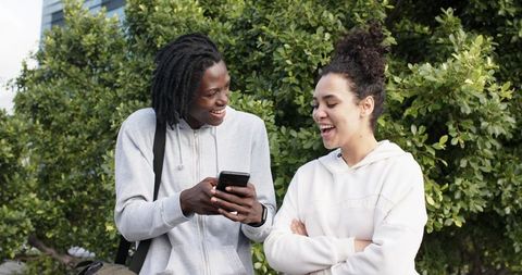 Multicultural Friends Laughing While Sharing Smartphone Outdoors Near Urban Greenery