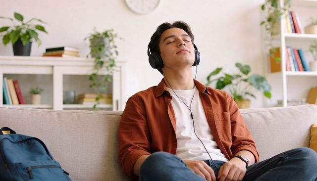 Young man relaxing on sofa listening to music with wired headphones in cozy living room