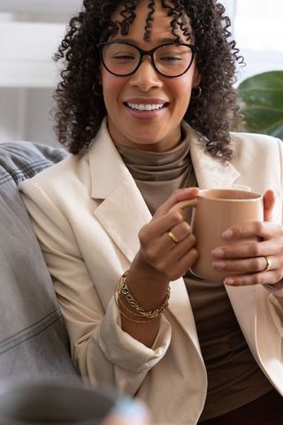 Smiling african american woman holding ceramic mug on sofa wearing blazer and glasses