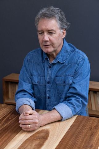 Senior Man in Denim Shirt Clasping Hands at Wooden Table