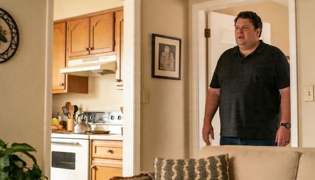 Standing middle-aged man surveying cozy kitchen from living room doorway, warm wood cabinets
