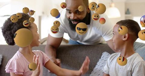 Smiling father playing with children in living room surrounded by floating emoji spheres