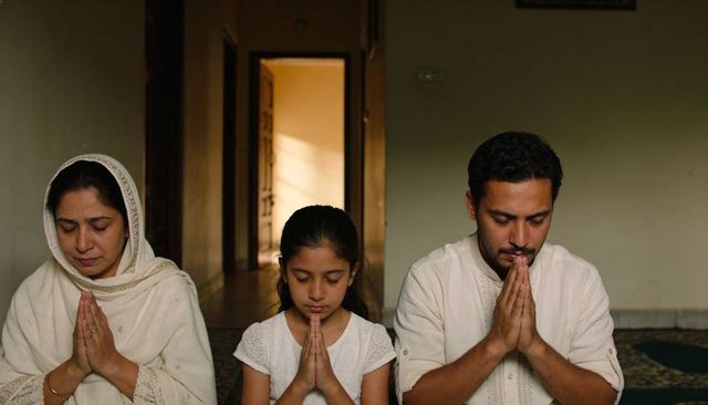 Muslim family praying together at home on patterned prayer rug with sunlit doorway