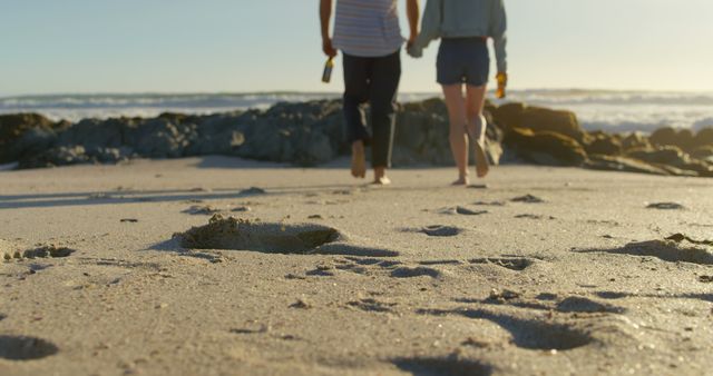 Couple holding hands with beer bottle walking on beach. Footprints in the sand 4k