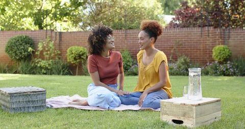 Mid-adult women chatting and sipping water on picnic blanket in sunny backyard garden