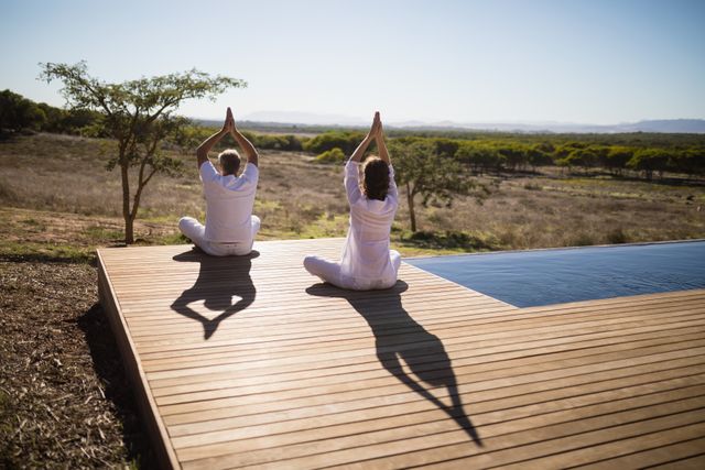 Couple practicing yoga on a wooden deck in a natural setting on a sunny day. Ideal for promoting wellness, mindfulness, and outdoor fitness activities. Suitable for use in health and wellness blogs, fitness websites, and lifestyle magazines.