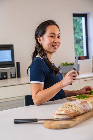 Asian woman preparing fresh bread and slicing green peppers on bright modern countertop