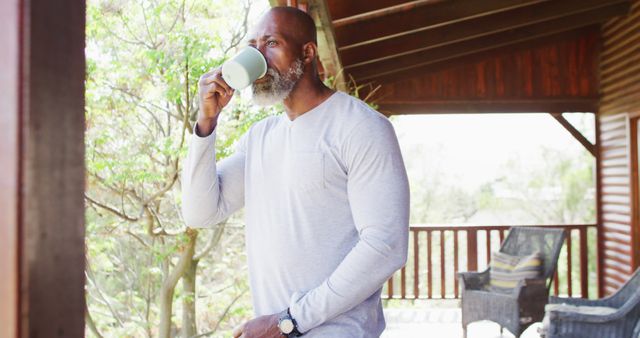 Mature man enjoying morning coffee on wooden balcony surrounded by nature. Wearing casual long sleeve shirt, appearing relaxed and peaceful. Ideal for use in lifestyle, wellness, or relaxation themed designs and blogs.