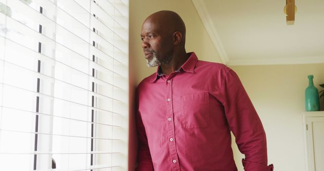 This image depicts a mature man standing by a window in a contemplative pose. He is wearing a casual red shirt and has a thoughtful expression as he gazes outside. Ideal for use in articles about mindfulness, elderly lifestyle, reflection, or personal stories. It can also be used in healthcare or retirement-related content to convey serenity or introspection.
