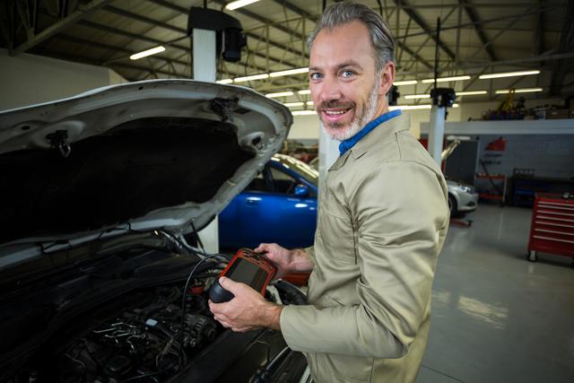 Mechanic using diagnostic tool in repair garage, smiling while working on car engine. Ideal for illustrating automotive services, car maintenance, and professional mechanic work. Useful for websites, brochures, and advertisements related to auto repair and vehicle diagnostics.