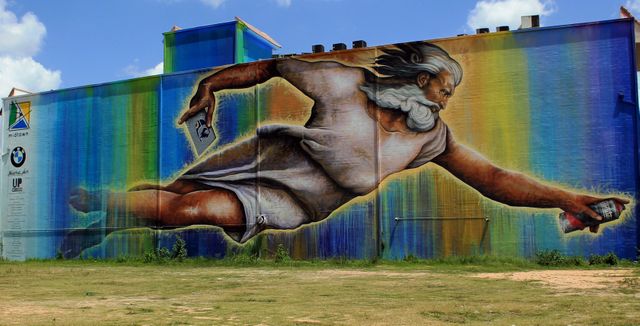 Colorful wall mural depicting a godlike figure reaching out, painted on the side of a building. The mural features vivid colors and a dynamic depiction of the figure, making it a vibrant piece of street art. This can be used for visual culture studies, urban art campaigns, public art appreciation, or promotional materials for art fairs and city tours highlighting modern and street art.