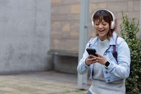 Young woman smiling, texting on smartphone with headphones and backpack in city courtyard