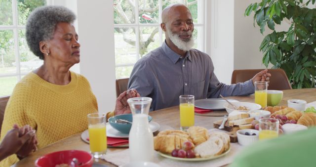 Senior African American couple displaying faith and togetherness while holding hands and praying before their breakfast. The setting provides a warm and inviting family atmosphere, ideal for concepts on gratitude, bonding, faith, and morning routines. Can be used for religious themes, family advertisements, senior living, nutrition, and healthy lifestyle content.