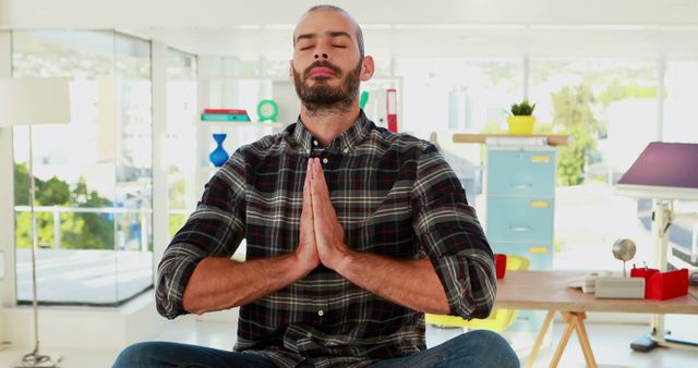 A middle-aged Caucasian man is practicing meditation in a bright office environment, with copy space. His serene expression and hands in a prayer position suggest a moment of mindfulness amidst a busy workday.