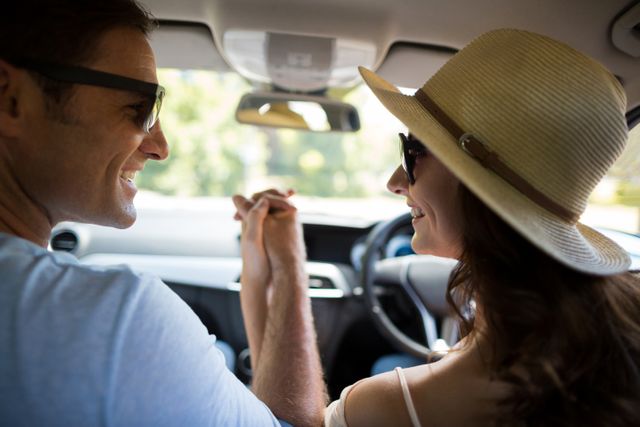 This image shows a happy couple holding hands while traveling in a car. They are both wearing sunglasses, and the woman is wearing a sun hat, indicating a summer road trip. This photo can be used for travel blogs, romantic getaway promotions, car advertisements, or articles about relationships and bonding.