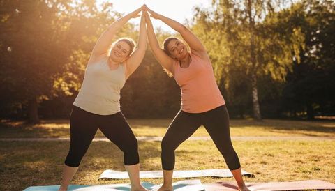 Two plus-size women doing partner side-bend yoga outdoors at golden hour on mats