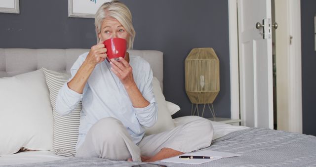 Senior woman sitting on bed in cozy bedroom, drinking coffee from red mug and enjoying a relaxed moment. Ideal for themes like retirement, comfortable living, morning routine, mental health, senior lifestyle, and home setting.