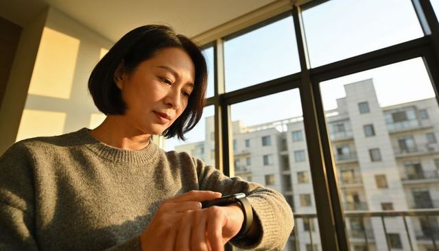 Middle-aged asian woman checking smartwatch at apartment window during golden hour