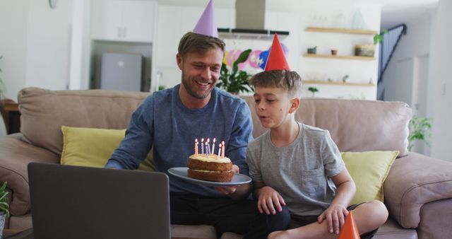 Caucasian man spending time with his son together, sitting on a couch, celebrating son's birthday, holding a birthday cake, using a laptop.