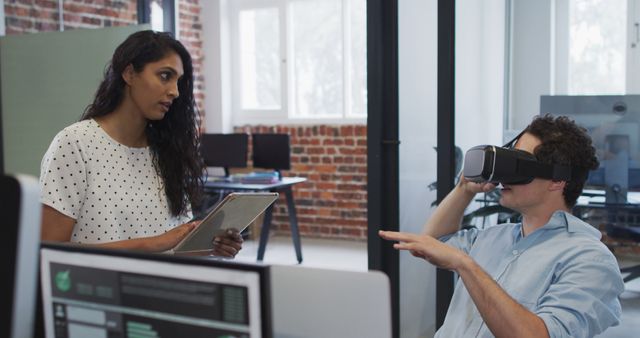 This image depicts a business setting in a modern office where a woman holds a digital tablet and a man interacts with a virtual reality headset. They are collaborating, possibly on a project or in brainstorming mode. Useful for concepts related to technology in business, innovation in the workplace, digital transformation, and modern work environments.