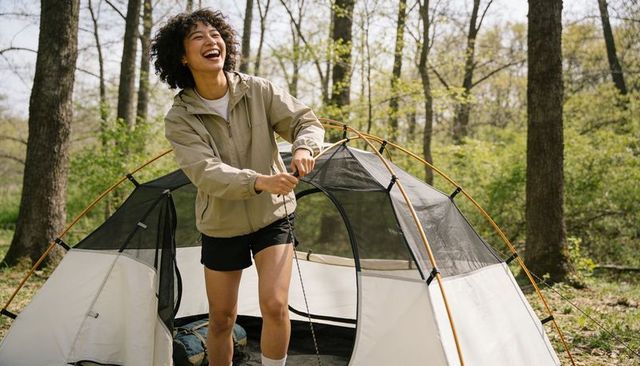 Woman laughing while pitching dome tent in sunlit forest campsite