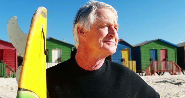 A senior Caucasian man stands on a sunny beach holding a surfboard, with copy space. His content expression and the vibrant beach huts in the background suggest a leisurely day spent surfing or enjoying the seaside.