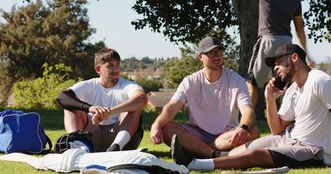 Male friends relaxing after cricket match sitting under tree on sunny park hill