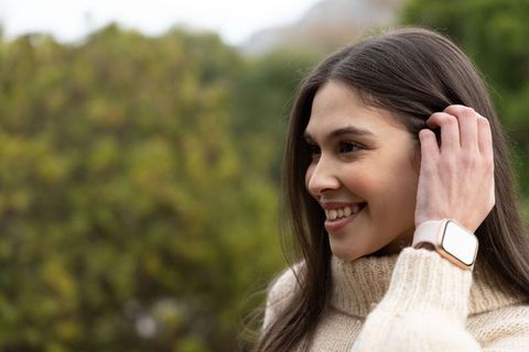 Smiling woman tucking hair wearing pink smartwatch and cream turtleneck casual outdoor portrait