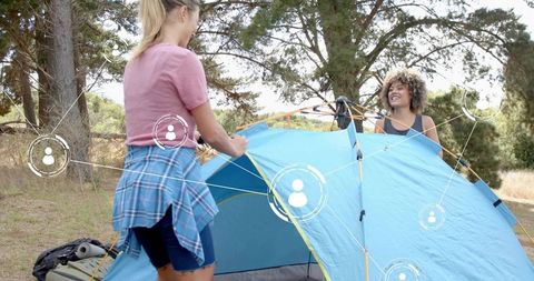 Friends pitching blue tent in sunlit pine forest demonstrating teamwork and outdoor fun