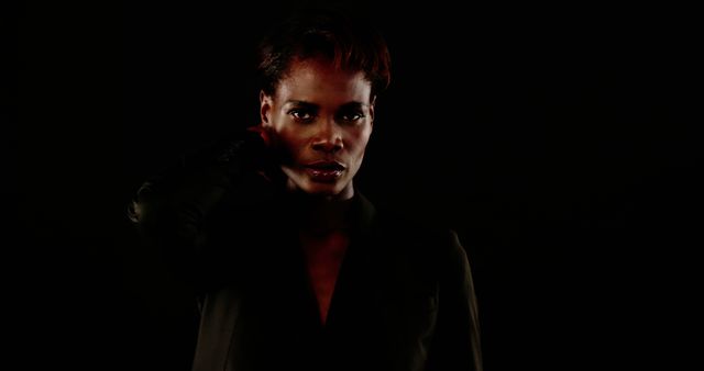 African American woman posing confidently in dark setting with black background. Great for themes of strength, mystery, professionalism, and fashion. Ideal for marketing, promotional materials, and blog posts focusing on empowerment and confidence.