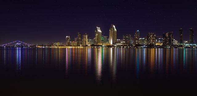 This image captures a stunning view of San Diego's skyline at night, with city lights beautifully reflecting on the calm ocean water. The high-rise buildings and illuminated bridge add to the urban appeal. Perfect for use in travel brochures, cityscape prints, websites related to tourism or real estate, and articles showcasing vibrant city life.