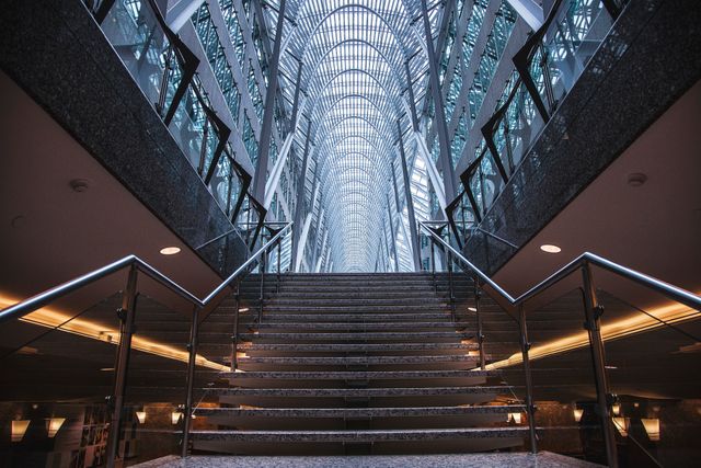 Impressive modern interior showcasing symmetrical design with expansive glass ceiling and prominent staircase. Steel structures add a futuristic touch, perfect for use in architecture, construction projects, corporate settings, or showcasing contemporary design themes.