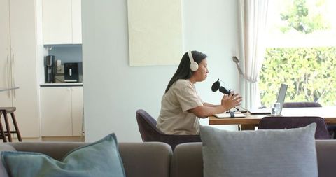 Mature woman recording podcast at home office dining table with laptop and microphone