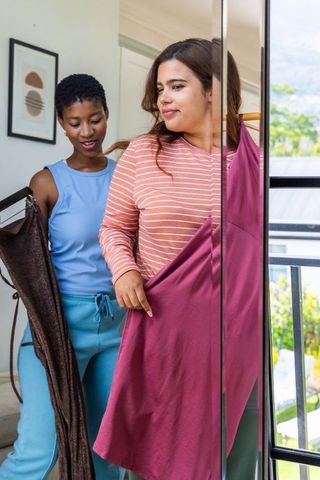 Two friends trying on dresses at home mirror, african american friend helping choose magenta dress