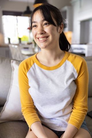 Smiling asian woman relaxing on beige couch in bright modern living room, cozy lifestyle