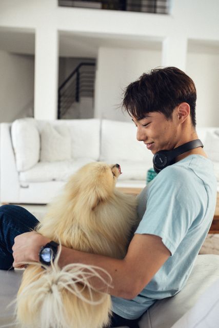 Asian teenage boy playing with his pet dog on couch in living room at home, copy space. Unaltered, pet owner, lifestyle, enjoyment, leisure activity.