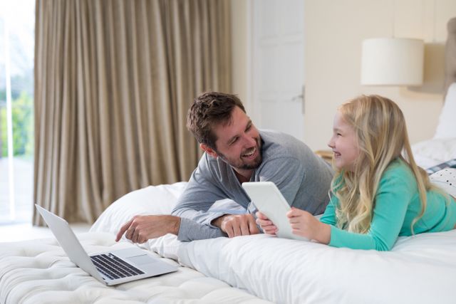 Father and daughter lying on bed using laptop and tablet, smiling and bonding. Perfect for family, technology, and parenting themes. Ideal for articles, blogs, and advertisements focusing on family life, digital devices, and home relaxation.