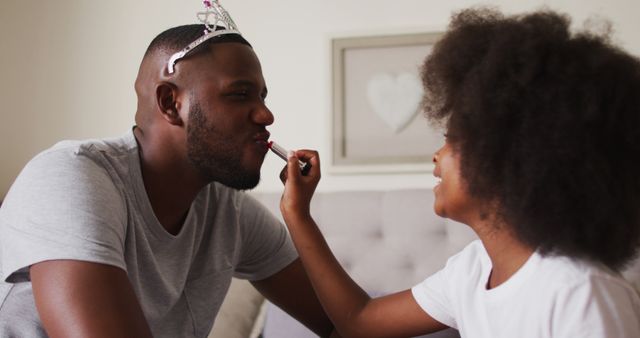 African american girl applying lipstick to her father father wearing crown at home. fatherhood parenthood concept.
