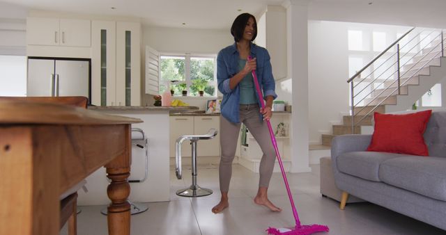 Young woman cleaning floor with a pink mop in a modern, tidy home. She wears casual clothing and looks content while performing household chores. Clean, modern interior features white cabinetry, island counter, and open staircase. Perfect for illustrations on domestic chores, home cleaning services, lifestyle blogs, or advertising cleaning products.
