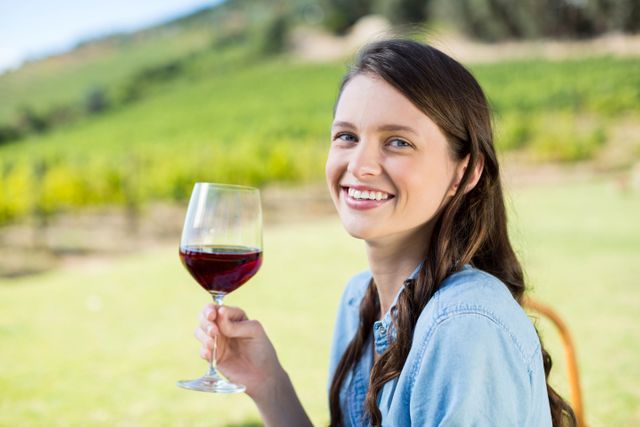This image shows a smiling woman holding a glass of red wine at a vineyard. Ideal for use in advertisements for wine brands, vineyard tours, lifestyle blogs, and promotional materials for outdoor leisure activities. It conveys a sense of relaxation, enjoyment, and connection with nature.
