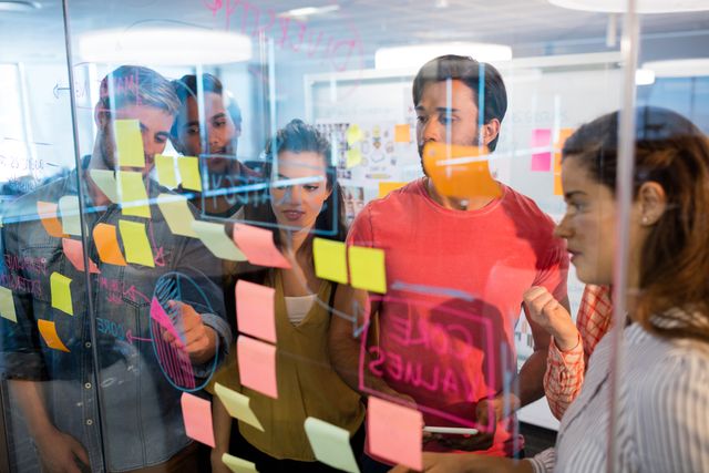 Group of young professionals collaborating and brainstorming ideas using colorful sticky notes on a glass window in a modern office. Ideal for illustrating teamwork, creative processes, project planning, and innovative business strategies.