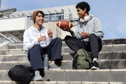 Multicultural college friends passing football on campus steps with backpacks, headphones