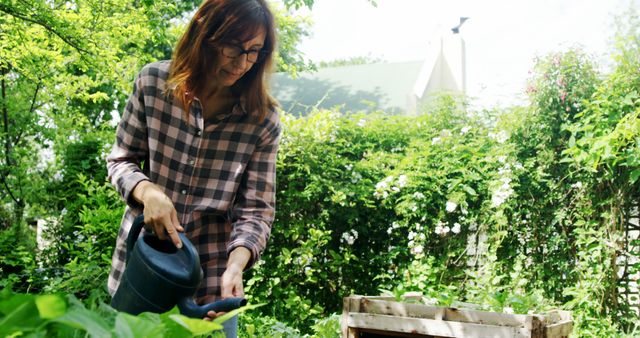Woman wearing checkered shirt watering plants in a lush green backyard. This image can be used for articles or advertisements about gardening, healthy living, outdoor activities, and sustainable practices.