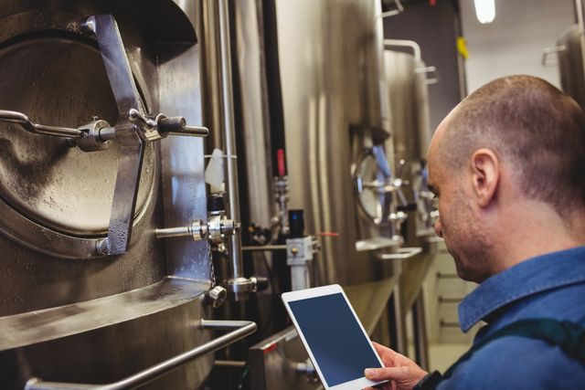 Brewery worker using digital tablet to inspect machinery. Ideal for illustrating modern brewing processes, industrial technology, and quality control in manufacturing settings. Useful for articles on brewery operations, technological advancements in brewing, and industrial equipment maintenance.