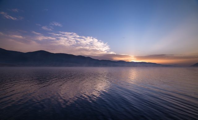 Beautiful image of a serene sunrise over a tranquil lake with mountains in the distance. The sky is a gradient of soft hues as the first light of day breaks. Ideal for use in travel blogs, nature websites, meditation apps, or relaxation backgrounds. Great for promoting themes of peace, tranquility, and natural beauty.